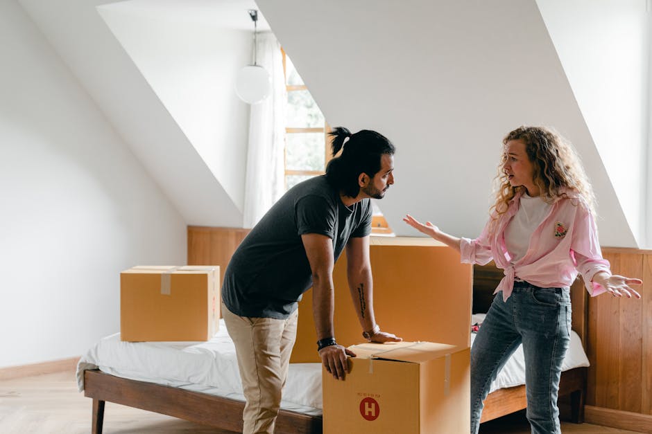 A man with dark hair tied back, wearing a black t-shirt and beige trousers, is seen inside a room with sloped white walls and wooden paneling. He is leaning slightly forward, resting hands on a cardboard box labeled with a red H, which is part of a home relocation process. Nearby, a woman with curly blonde hair, dressed in a pink top with rolled-up sleeves and blue jeans, is engaged in a discussion, gesturing with both hands as if explaining or instructing. The room is illuminated by natural light coming through a window with white curtains, and the background shows a bed with a white mattress and another closed cardboard box placed on it. Several other boxes and packing materials are visible, suggesting an active moving or packing session, possibly coordinated by a professional removals service such as Man and a Van Croydon. The setting emphasizes packing and moving logistics within a residential space, aligned with house removals services related to the Thornton Heath relocation guide.