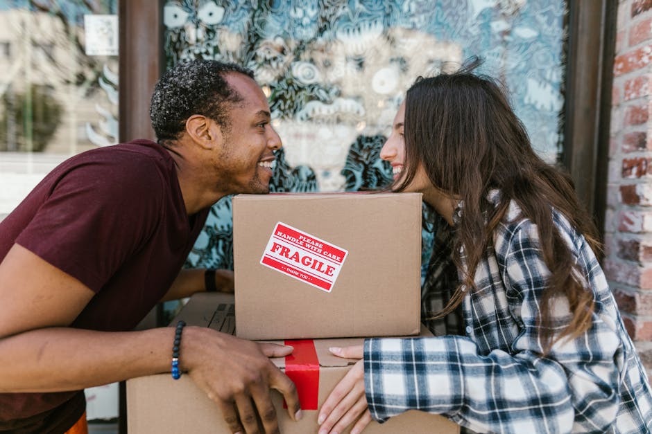 A young man and woman are engaged in a house removal process outside a building with a large window and brick wall. The man, wearing a maroon T-shirt and a bracelet, is smiling as he hands over a medium-sized cardboard box marked with a red 'Fragile' sticker to the woman, who is dressed in a plaid shirt. They are positioned close together, with the woman holding the box while smiling and looking at the man. In front of them, there are multiple boxes stacked, some wrapped with red tape, indicating packing and moving preparation. The background shows a glass window with decorative designs and a slightly blurry outdoor setting, suggesting daylight. This scene captures the process of packing and loading household items, illustrating home relocation activities often handled by [COMPANY_NAME] during a furniture transport or home removal service.