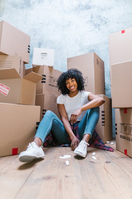 A smiling woman stands indoors against a blue textured wall, holding an open cardboard box labeled 'GLASS' in red letters, indicating careful packing during a house removal or home relocation. She is 