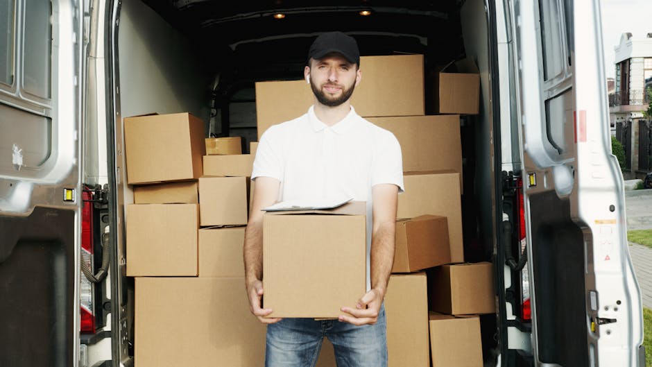 A man standing at the rear of a large van during a home relocation, holding a medium-sized cardboard box with both hands. The van is parked outdoors on a paved area near residential buildings, and the interior of the van is loaded with numerous stacked cardboard boxes of various sizes, some wrapped in clear plastic and others exposed. The boxes are arranged neatly, filling the space from the floor to the roof of the vehicle. The man is dressed in casual clothing, including a black cap, a white polo shirt, and jeans. He is smiling slightly and appears to be in the process of loading or unloading moving boxes as part of furniture transport and packing and moving services. The environment is well-lit, with natural daylight illuminating the scene, illustrating the logistics involved in a typical house removal.