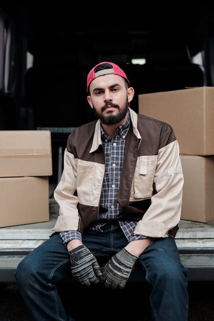 A young man with a beard, wearing a red cap backwards, a plaid shirt, and a brown and beige jacket, is sitting on the loading ramp of a van used for home relocation. The van's interior is dark, with the back doors open, revealing several cardboard boxes of various sizes, some stacked on top of each other, positioned both inside the vehicle and on the pavement behind him. The man is wearing grey and black striped gloves, indicating he has been involved in packing or handling items for moving. He appears to be taking a break during the furniture transport or packing process, which is part of the house removal services offered by Man and a Van Croydon. The scene is set outdoors, with natural lighting highlighting the boxes and the man, emphasizing the logistics involved in furniture transport and moving services in the Croydon area, specifically for CR0 postcode moves.